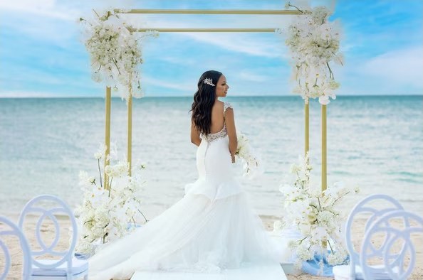 Novia de espaldas frente a un arco dorado con flores blancas junto al mar